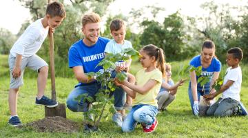 Image shows a male and female volunteer in blue volunteer t-shirts. They are kneeling down to help four young people plant trees. 