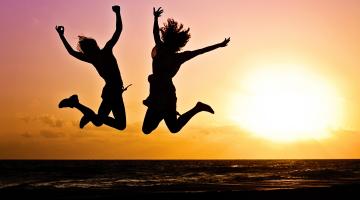 Images shows the silhouette of two young people jumping in the air against a sunset background 