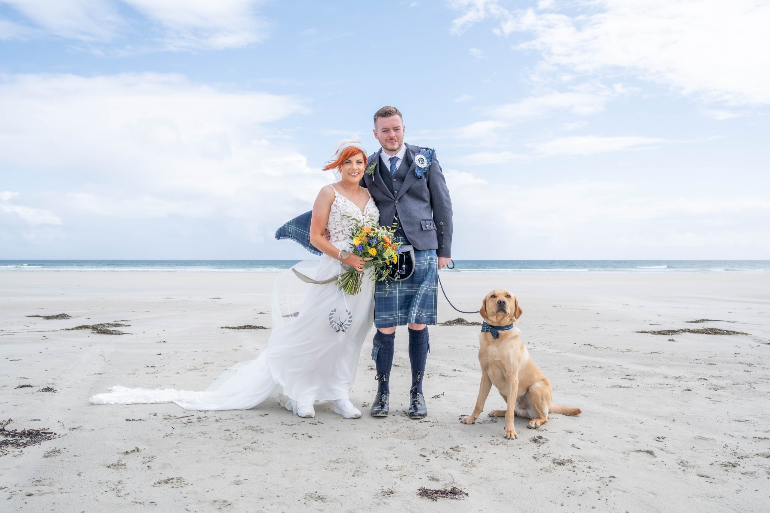 woman in white dress with a man in a kilt and a dog on the beach