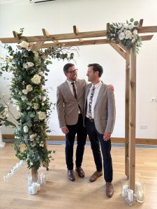 2 men in suits under a wooden arch with flowers