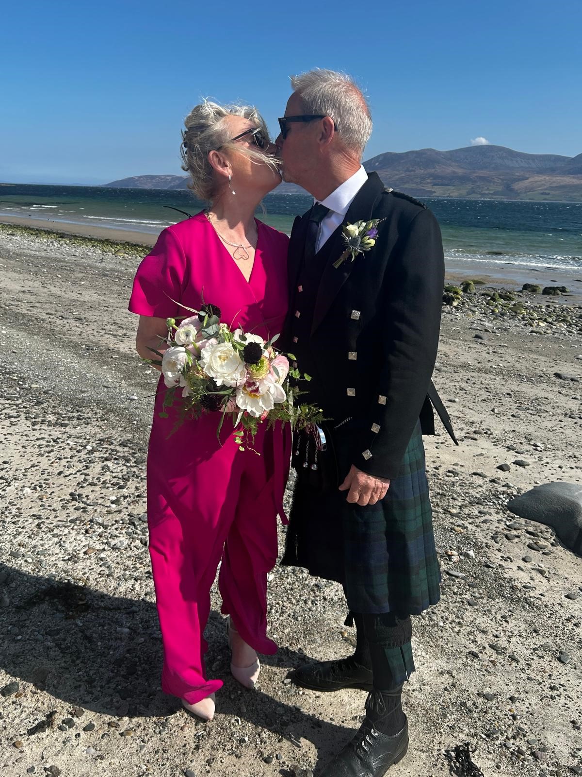 a man in a kilt hugging a woman in pink dress with flowers in her hand on a beach