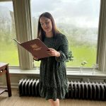 girl in a dress standing in front of a window reading a book