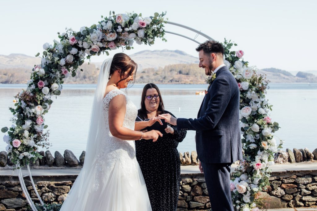 one woman in white dress holding man in suit hand. Woman in middle with flower arch behind