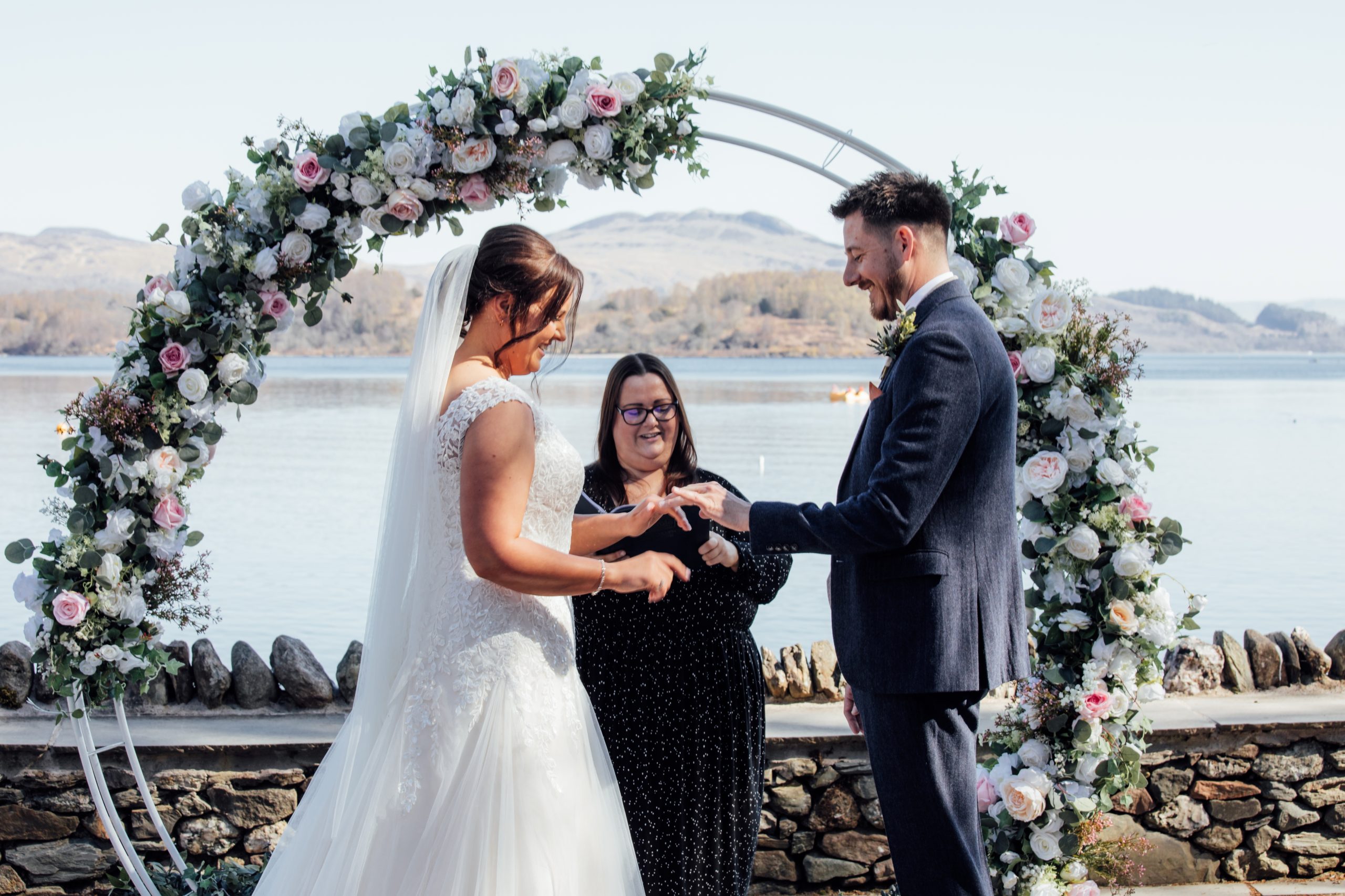 one woman in white dress holding man in suit hand. Woman in middle with flower arch behind