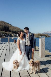 Bride in white dress and man in suit on a pier holding leads for a dog each