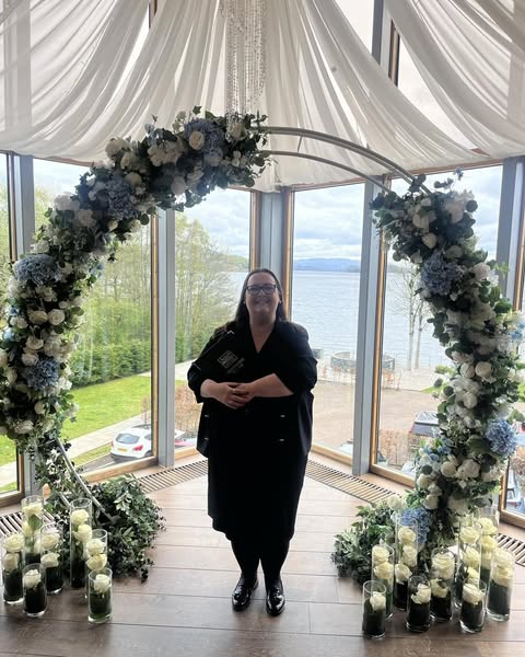 a woman in black dress in a room standing in front of a half moon arch with flowers round it