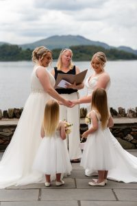 3 woman standing 2 in white dresses with water and hills behind