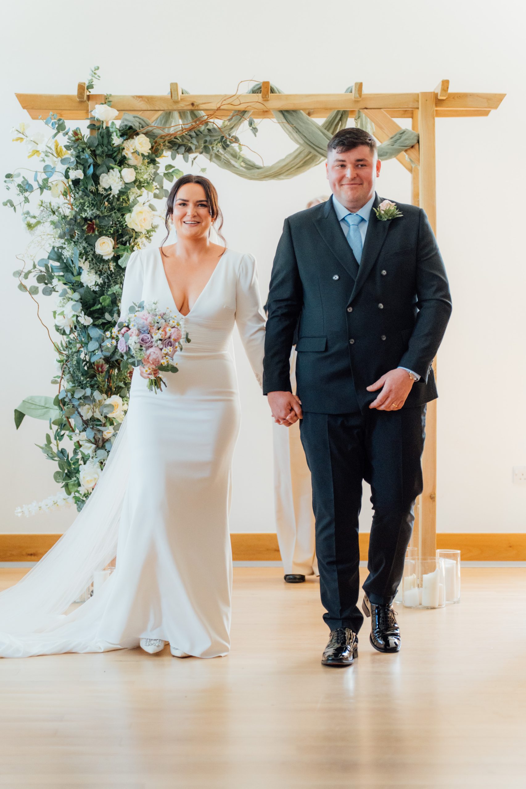 man in suit handling woman in brides dress with flower arch behind them