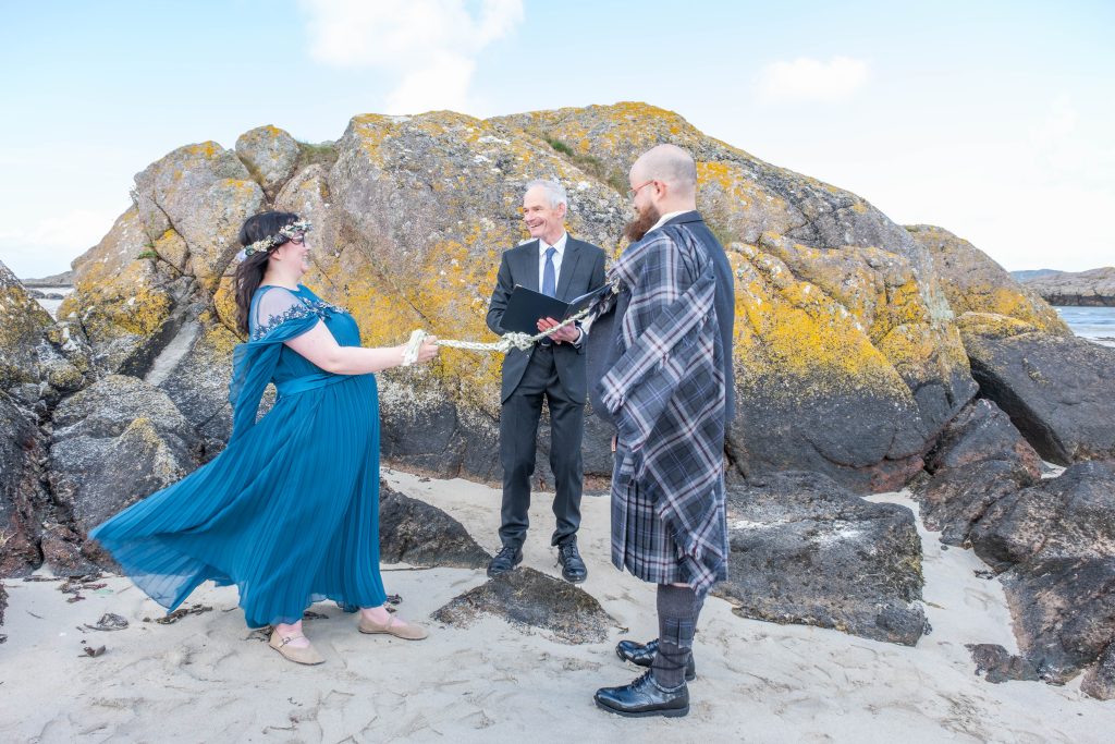woman in blue dress holding cord with a man in a kilt. Man in suit between them background is beach big rocks