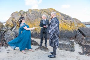 woman in blue dress holding cord with a man in a kilt. Man in suit between them background is beach big rocks