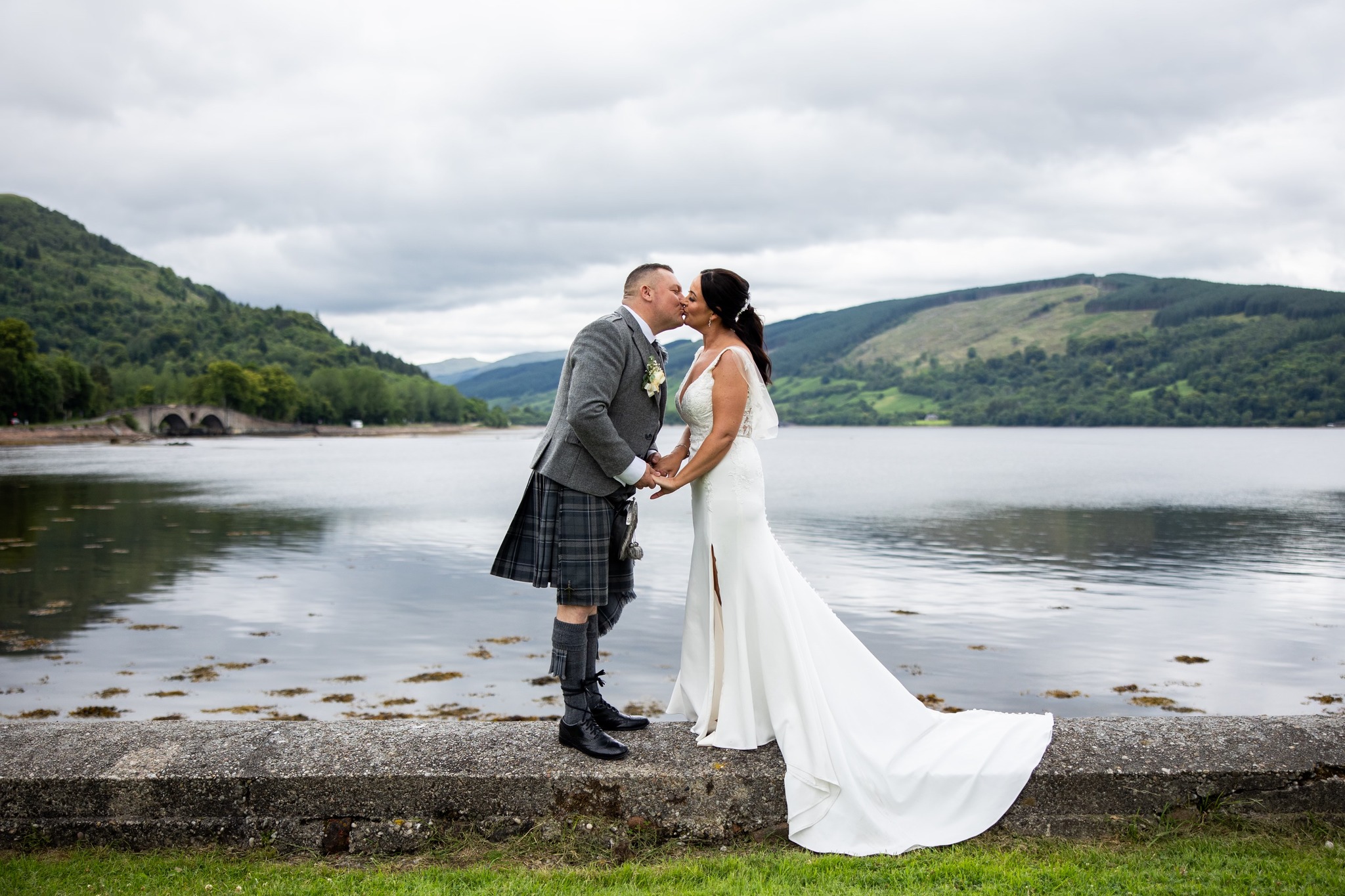 man in a kilt and a woman in a white dress kissing with mountains and water in the background