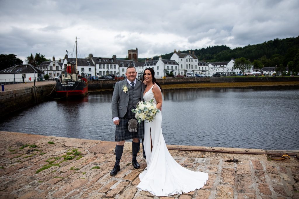 man in a kilt and a woman in a white dress with shops and water in the background