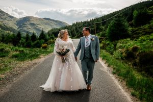 woman in brides dress holding hands with man in 3 piece suit in the middle of a road with mountain around them
