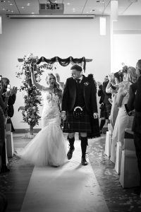 Black and white photo - woman in white dress hand in air walking with Man in kilt