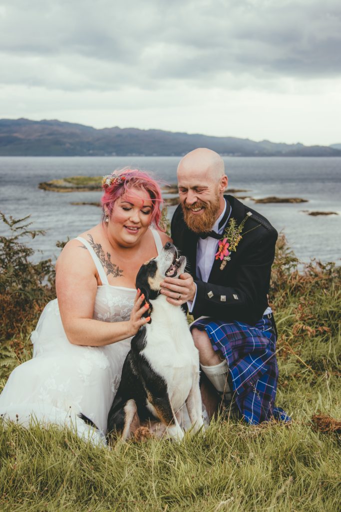 woman with pink hair in white dress bending down with man in a blue kilt looking at a dog