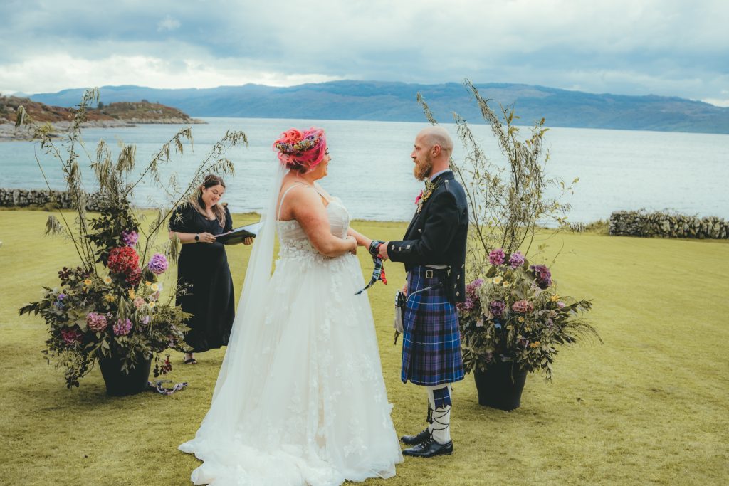 Woman in white dress holding hands with man in Kilt on grass water in the background