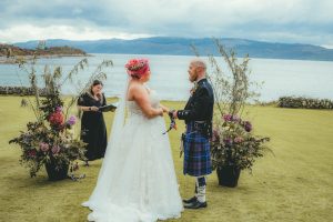Woman in white dress holding hands with man in Kilt on grass water in the background