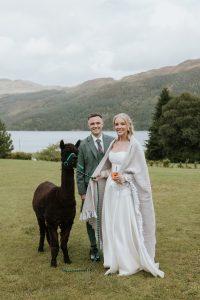 woman in white dress standing with man in green kilt on grass next to an alpaca