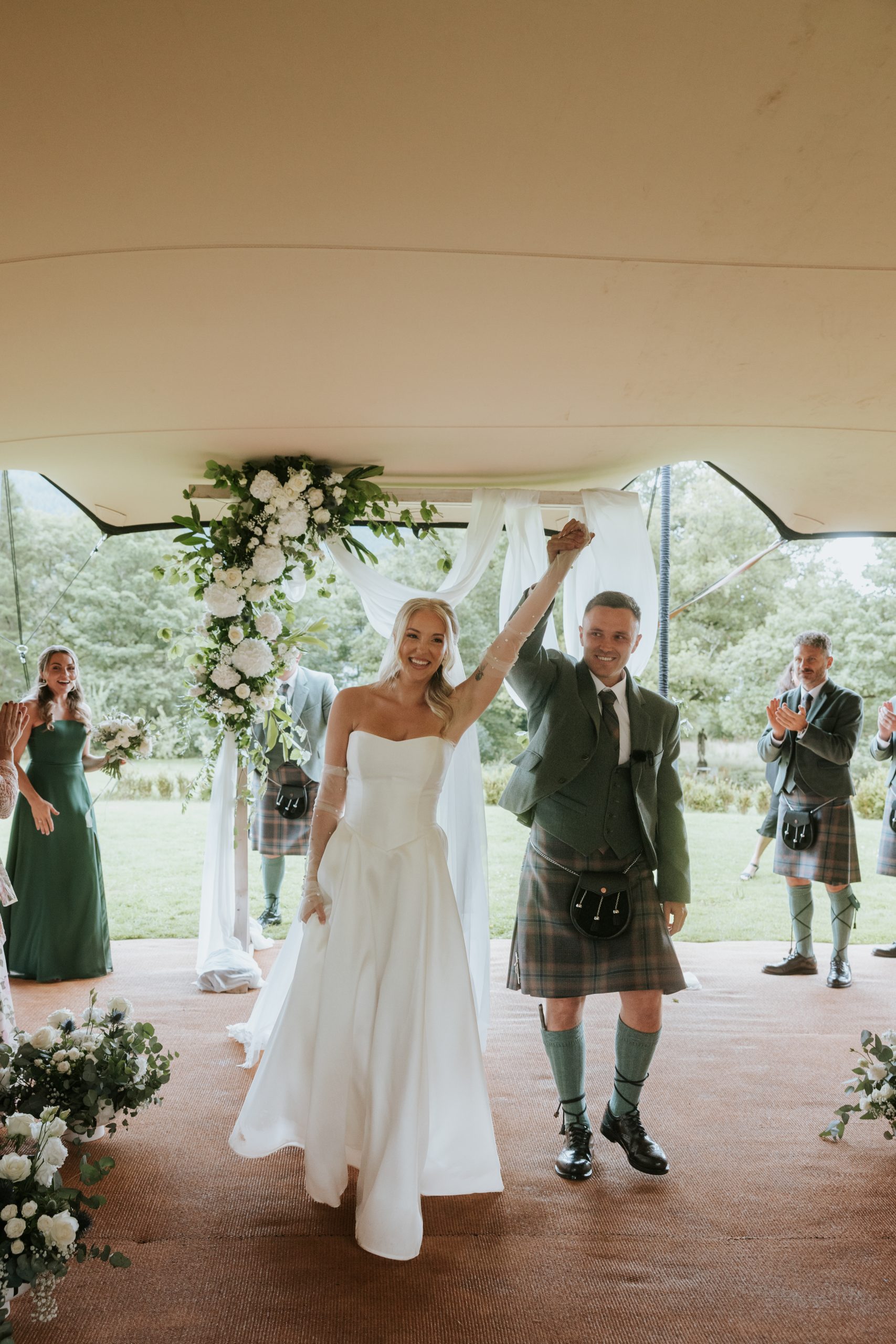 woman in white dress holding arm up in air with man in a green kilt