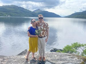 Lady in a blue top yellow skirt holding wild flowers standing with man water and mountains in the background