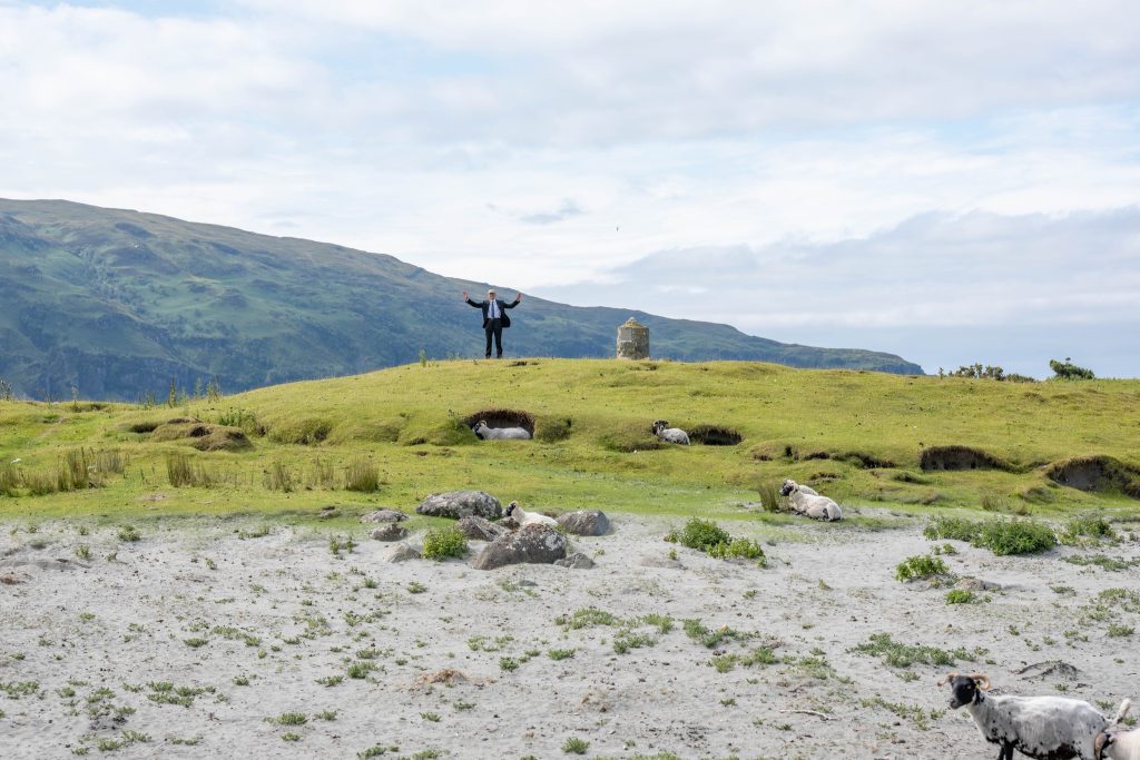Man in a suit standing on top of a hill surrounded by grass and sheep