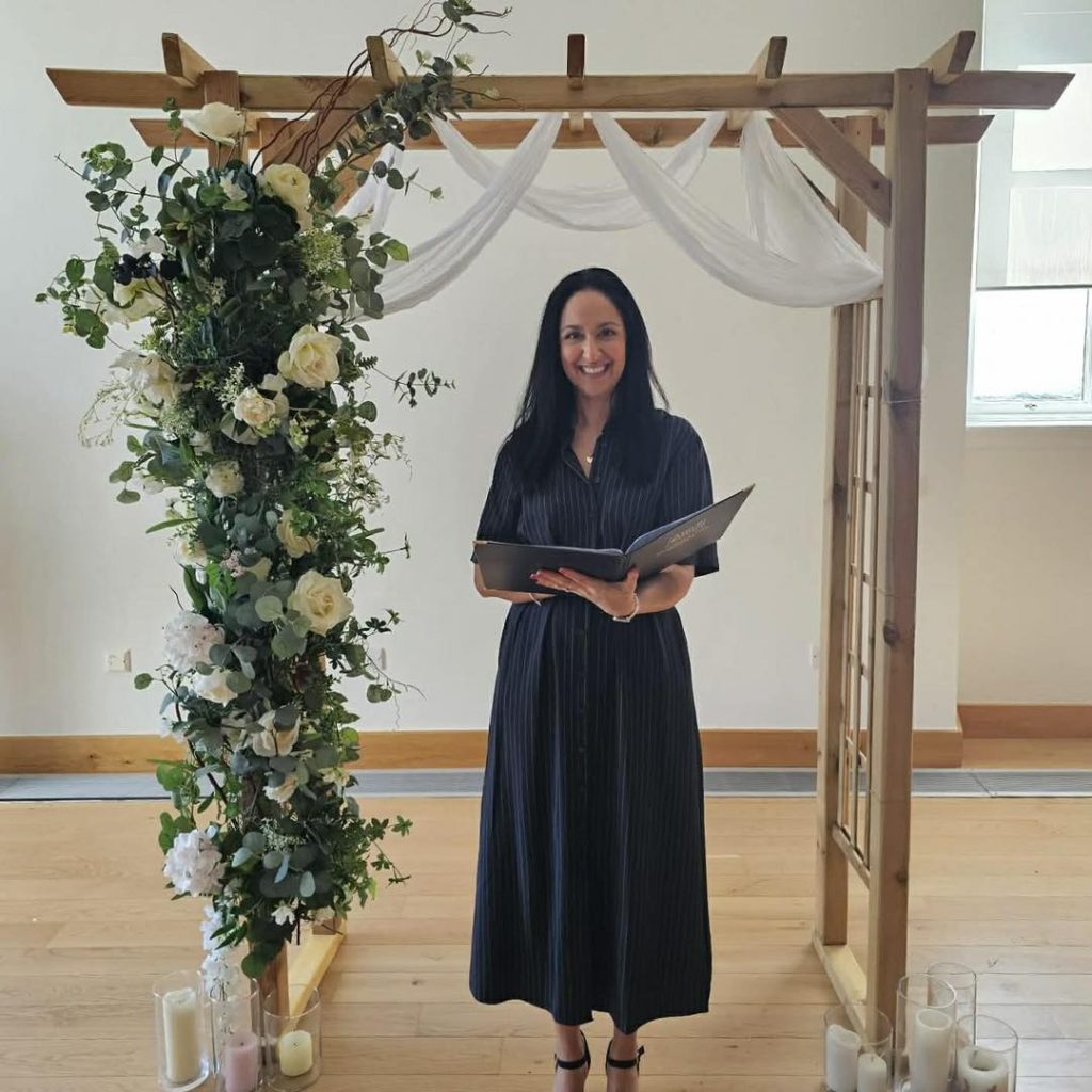 A woman in a black dress with a book under a wooden arch with flowers down one side