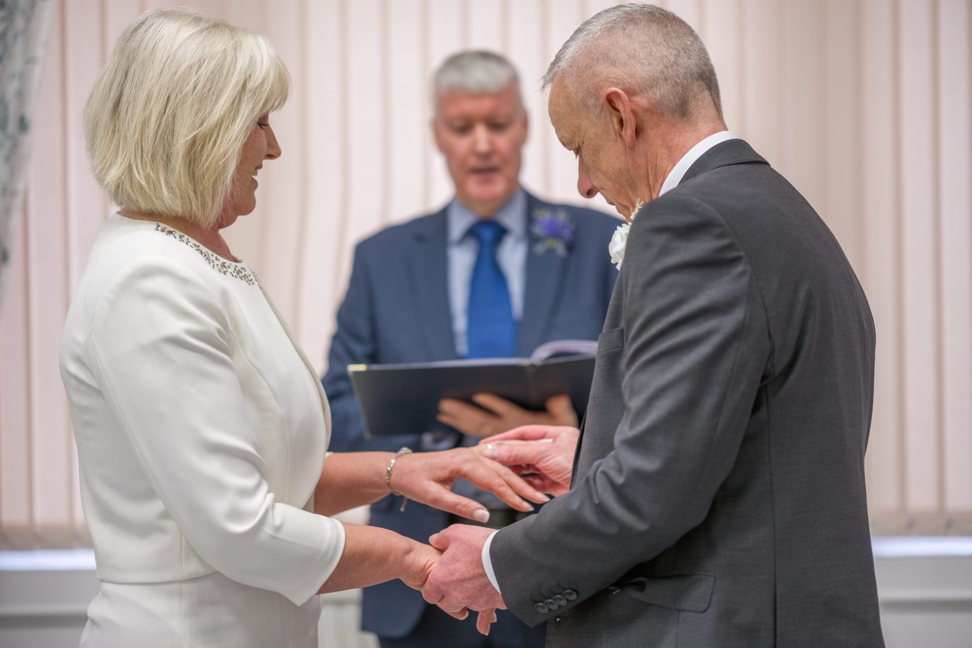 A man in grey suit standing holding hands with a woman in white suit and a man in the middle of them wearing a suit holding a book