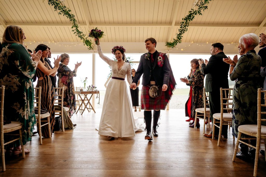 A lady in white holding up arm with flowers in her hand and holding the hand of a man in a kilt