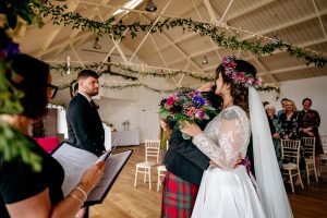 Man in red kilt hugging a woman in white dress with flowers in her hand and on her head.