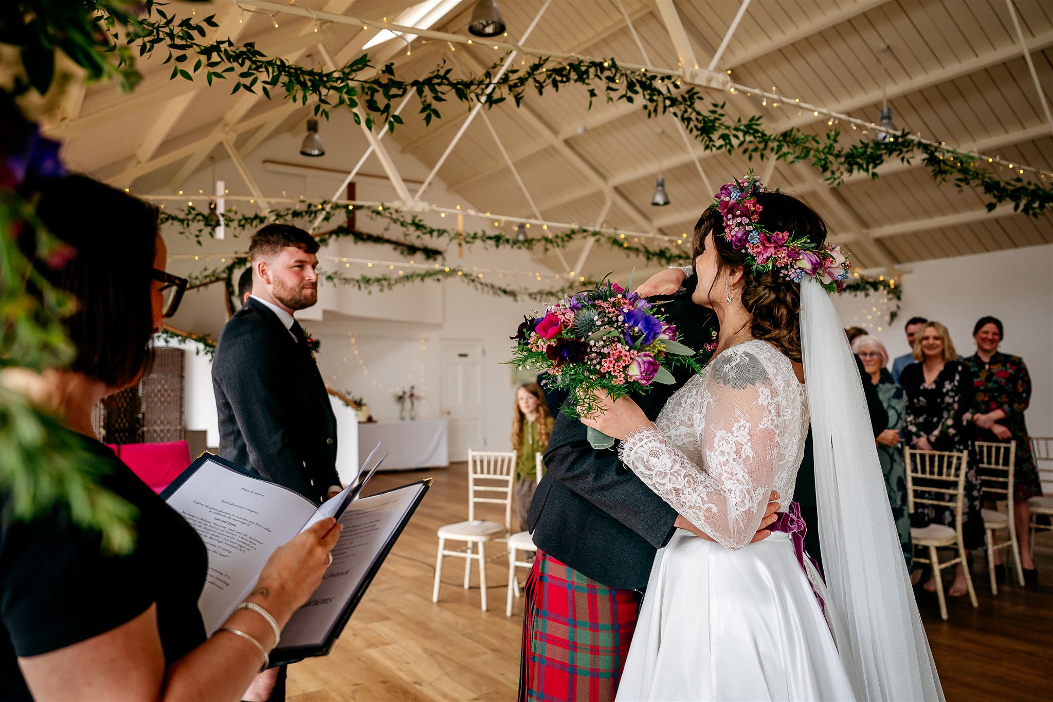 Man in red kilt hugging a woman in white dress with flowers in her hand and on her head.