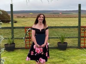 woman in a black dress with pink flowers at the bottom standing smiling in a field