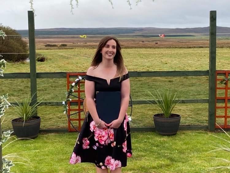 woman in a black dress with pink flowers at the bottom standing smiling in a field
