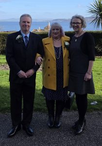 A man in a suit linked arm with a woman with dotty dress and a yellow jacket on next to another woman in a black dress.
