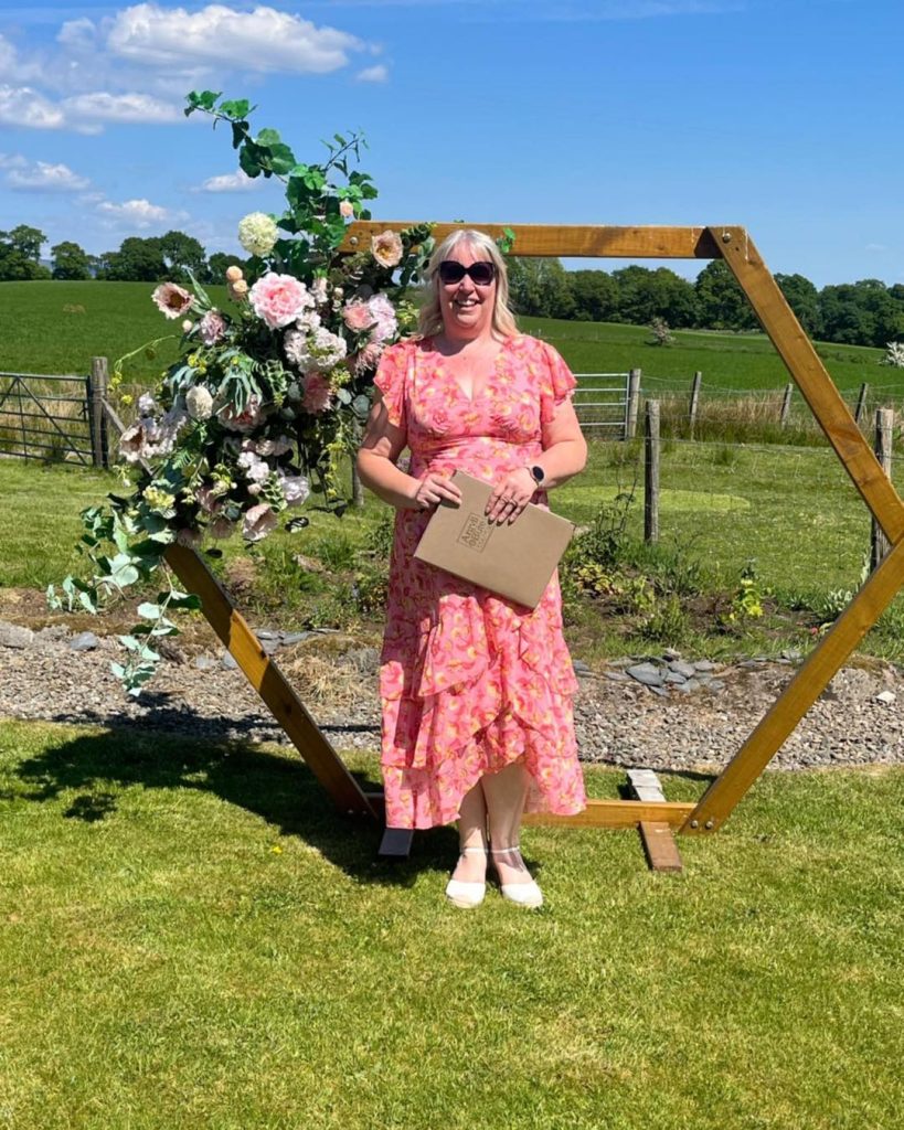 Blonde woman with sunglasses on wearing a flowery pink dress standing in front of a wooden hexagon arch