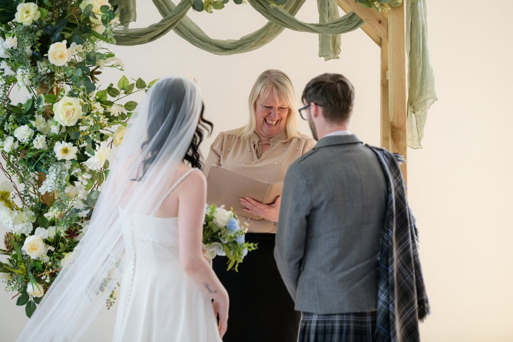 Blonde woman laughing under a wooden arch with a bride and a groom in a kilt in front of her