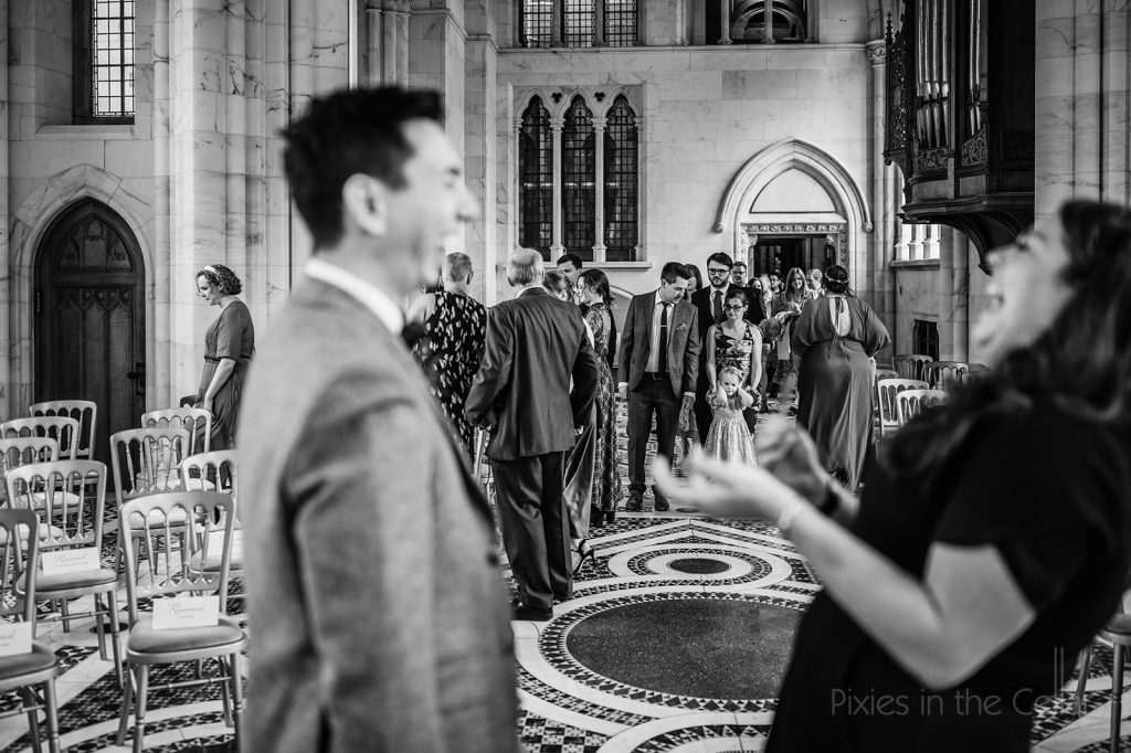 Black and white photo in marble chapel people grouped together at the back and a man and woman laughing