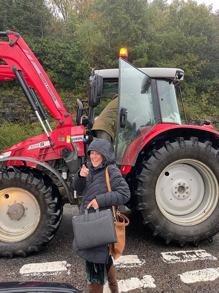 Woman with black jacket standing pointing figure holding black bag next to a tractor