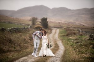 A man in a suit kissing a woman in a wedding dress while walking up a outdoor farm track