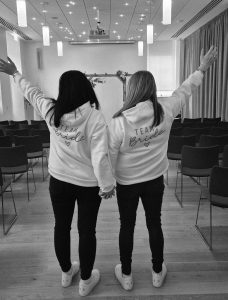 2 woman holding holding hands with their other arm in the air. Both wearing a hoody with Team Bride on the back. Black and white photo