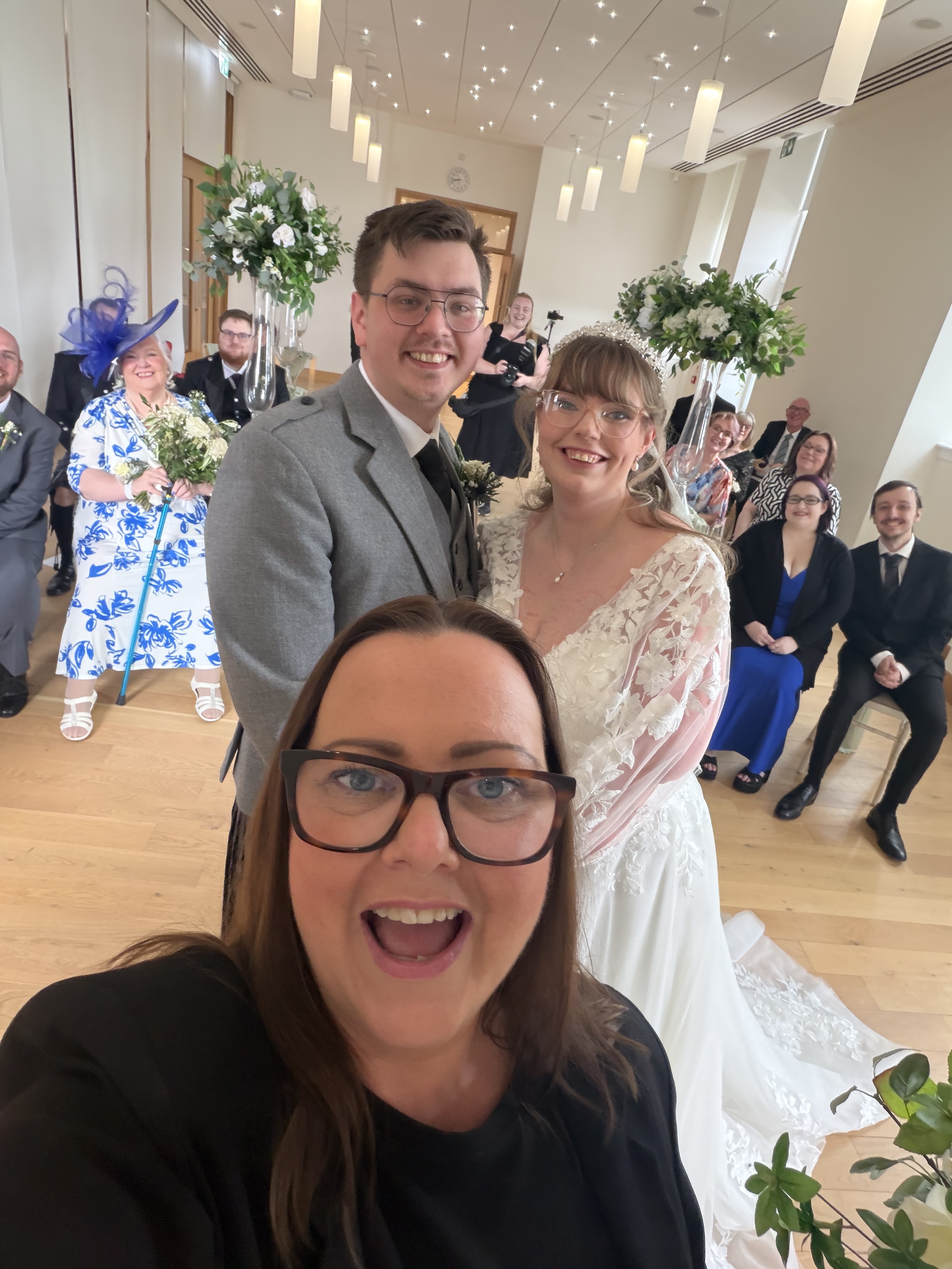 a marriage room filled with people and a woman in black taking a selfie with bride and groom