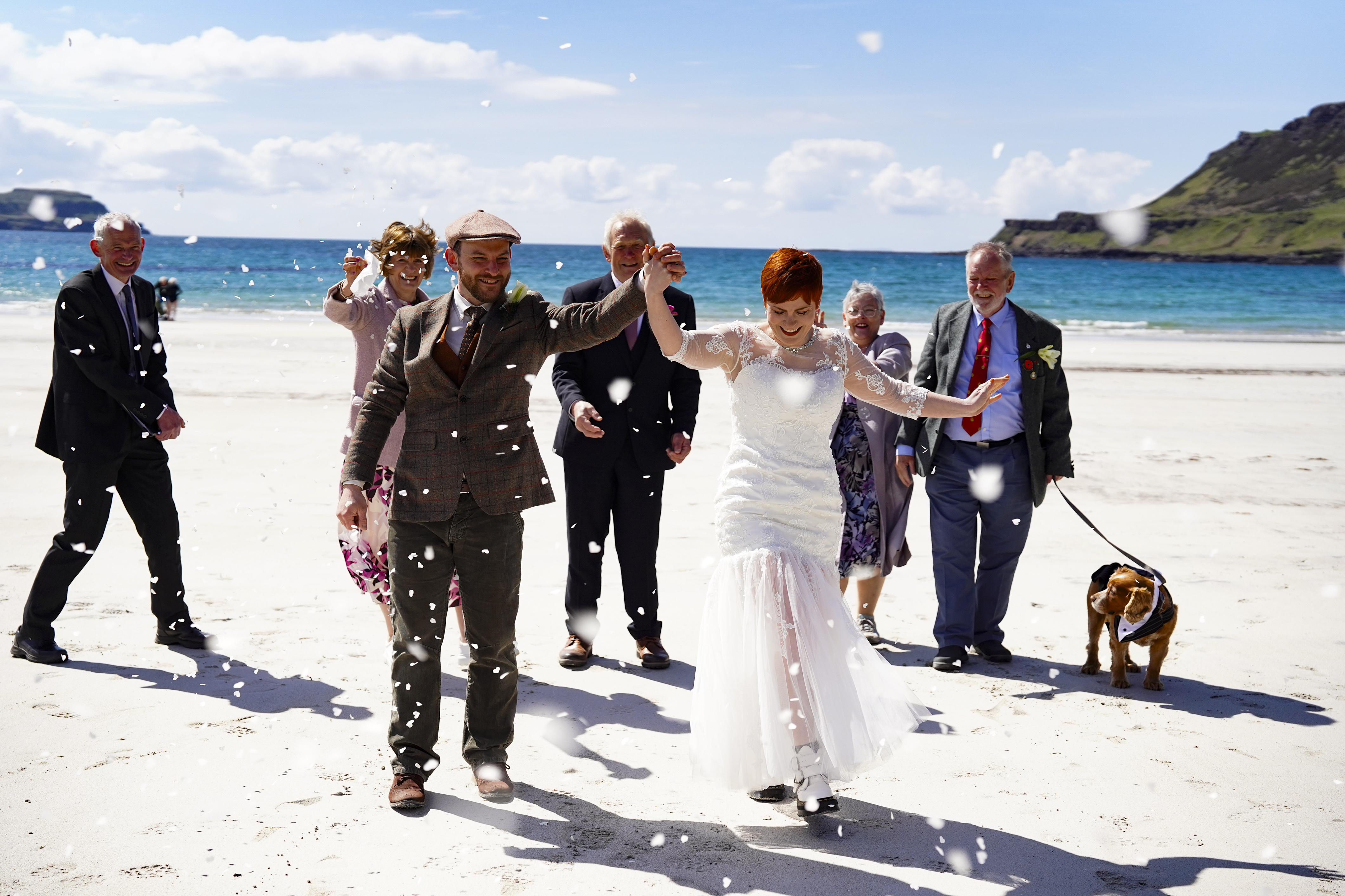 a group of people on a beach and in front is a groom and bride holding their arms up in the air