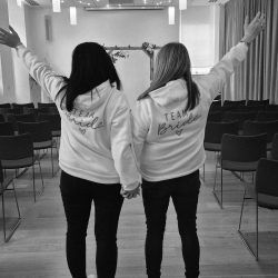 2 woman holding holding hands with their other arm in the air. Both wearing a hoody with Team Bride on the back. Black and white photo