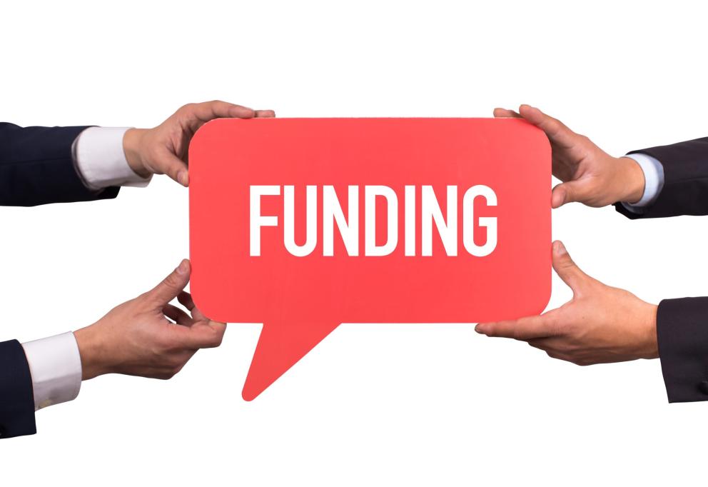 Images shows four hands holding a sign up with the words funding on a red background. 