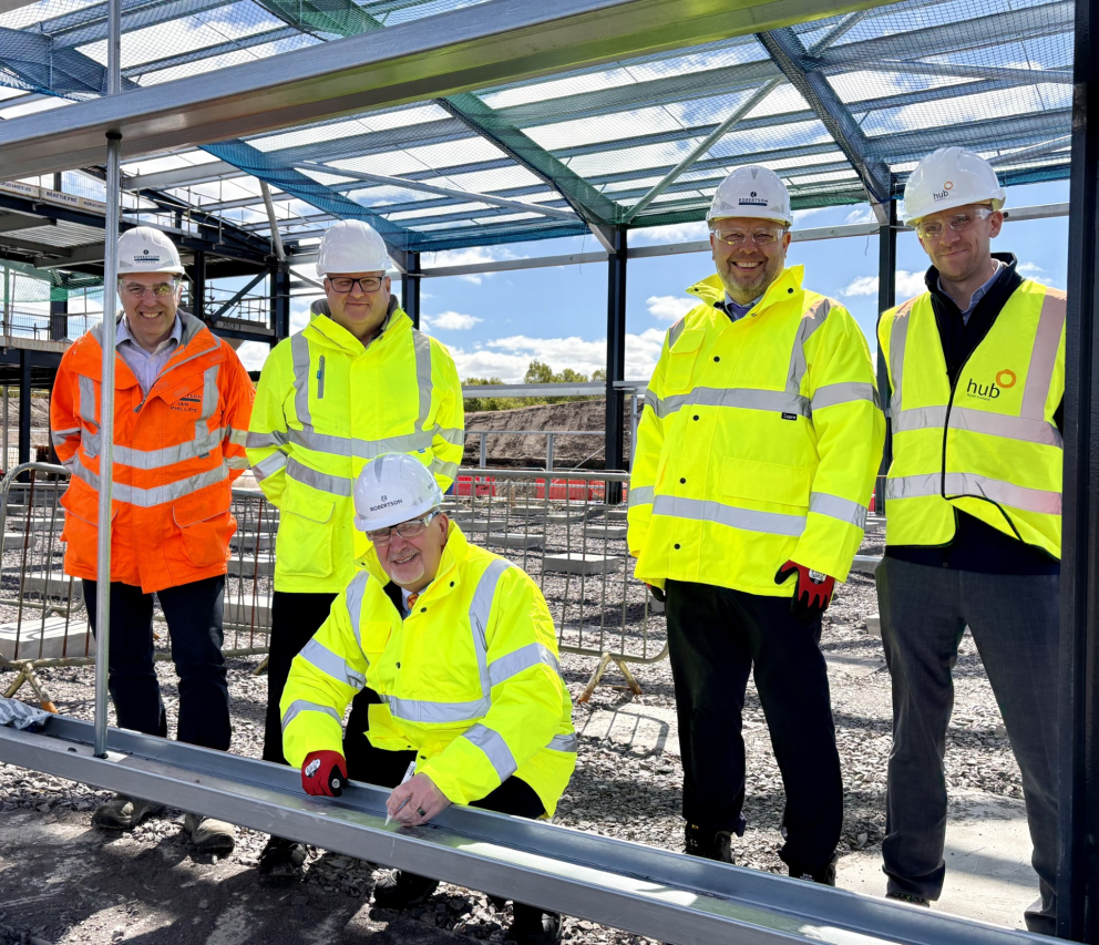 Five men, in Hi Vis jackets and hard hats stand on a building site underneath a steel frame of a building