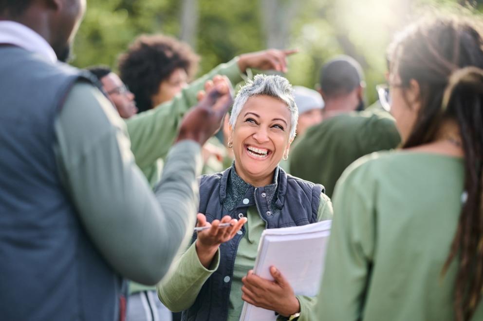Image shows a lady smiling holding a notepad talking to a group of people 