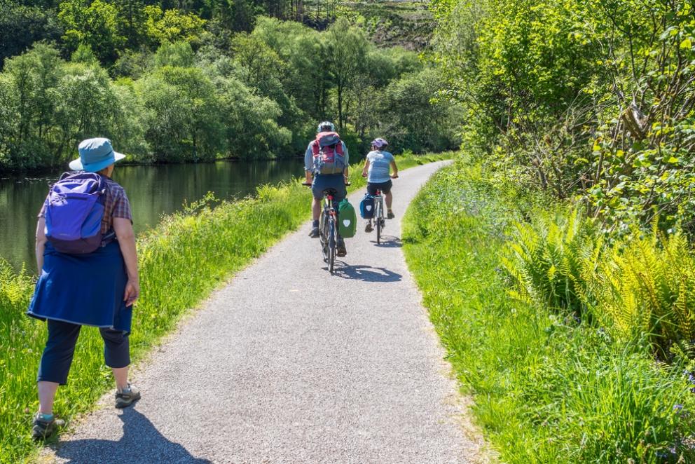 A woman walks along a path next to a river with two cyclists ahead of her