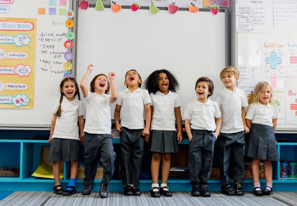 Image shows a line of primary school children in uniform standing in front of a whiteboard laughing 