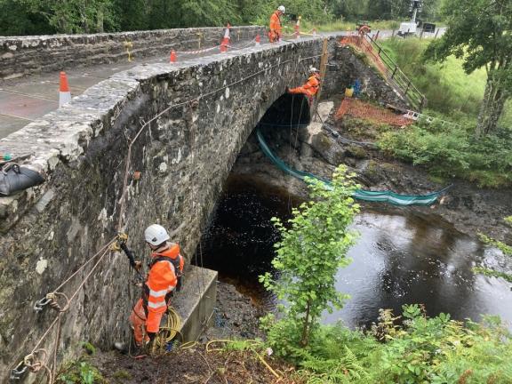 Workmen on bridge, one hangs from ropes over the side.