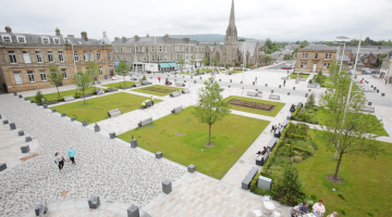 An aerial image of Helensburgh's Colquhoun Square. To the bottom right of the image, a group of people are sitting at a cafe table. 