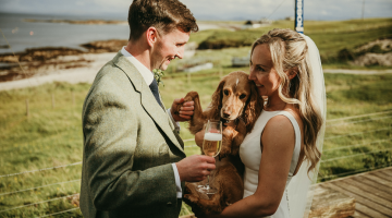 Photo of a bride and groom. The groom is on the left. The bride is holding a cocker spaniel.